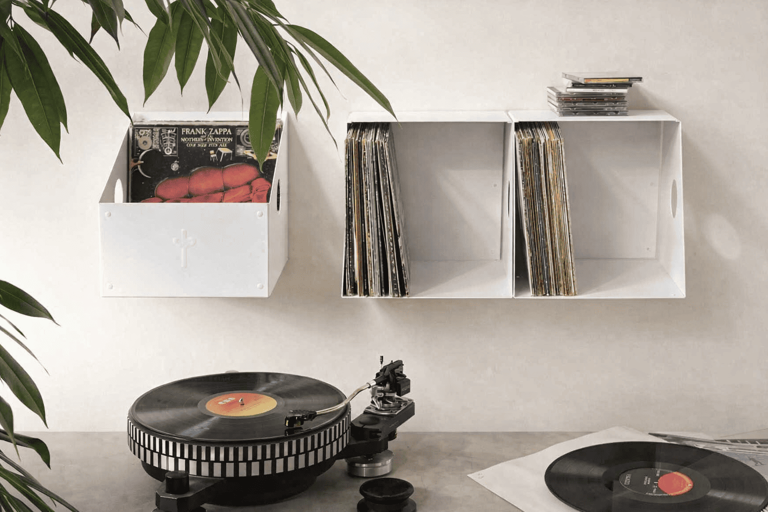 Dans un intérieur minimaliste et apaisant, un bac à vinyle blanc suspendu au mur accueille des disques soigneusement rangés au-dessus d’une platine noire. De grandes feuilles vertes encadrent la scène, tandis qu’une lumière douce souligne un coin musique élégant et chaleureux.