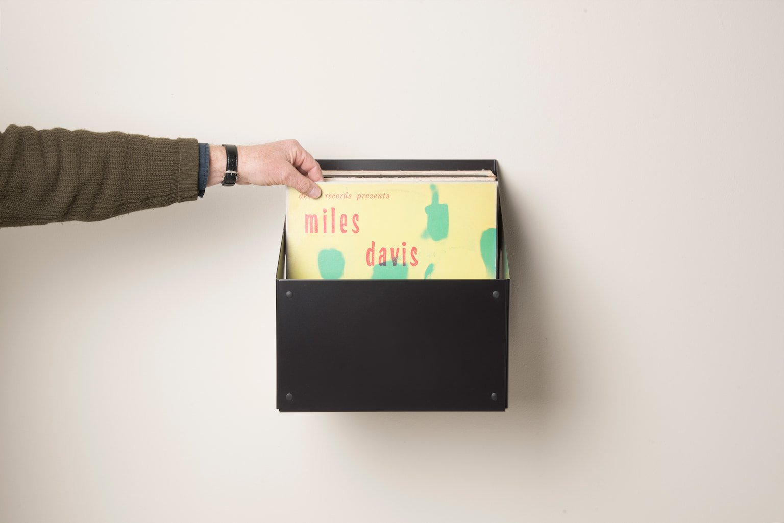Bac de rangement vinyle mural noir en métal contenant plusieurs disques 33 tours, avec une personne faisant glisser une pochette pour parcourir sa collection.