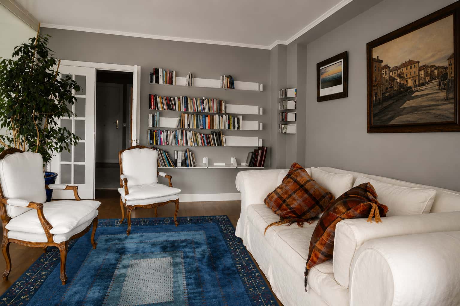Bibliothèque murale blanche sur mur gris, harmonisée avec le canapé et les fauteuils blancs du salon