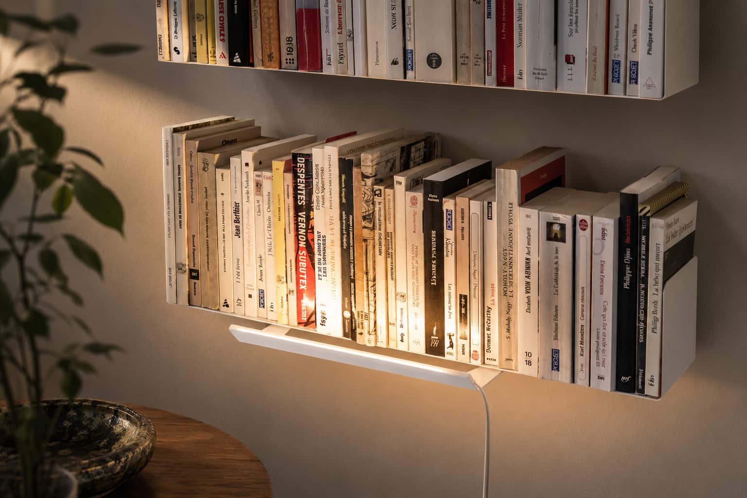 Bibliothèque murale blanche remplie de livres, mise en valeur par un éclairage chaud sous les étagères dans un intérieur contemporain.