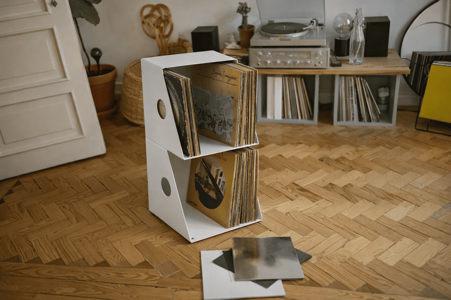 Boîte blanche moderne de rangement vinyle 33 tours posée au sol, remplie de disques vinyles, dans un salon chaleureux avec parquet et platine audio en arrière-plan.