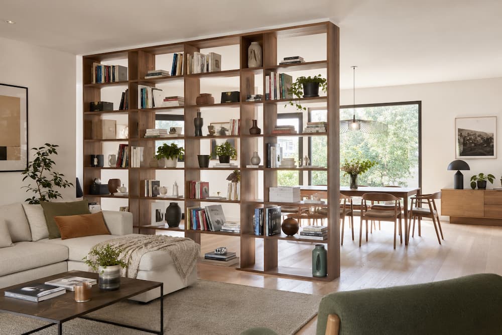 Salon et salle à manger séparés par une grande cloison-bibliothèque ajourée en bois, remplie de livres, plantes et objets décoratifs.