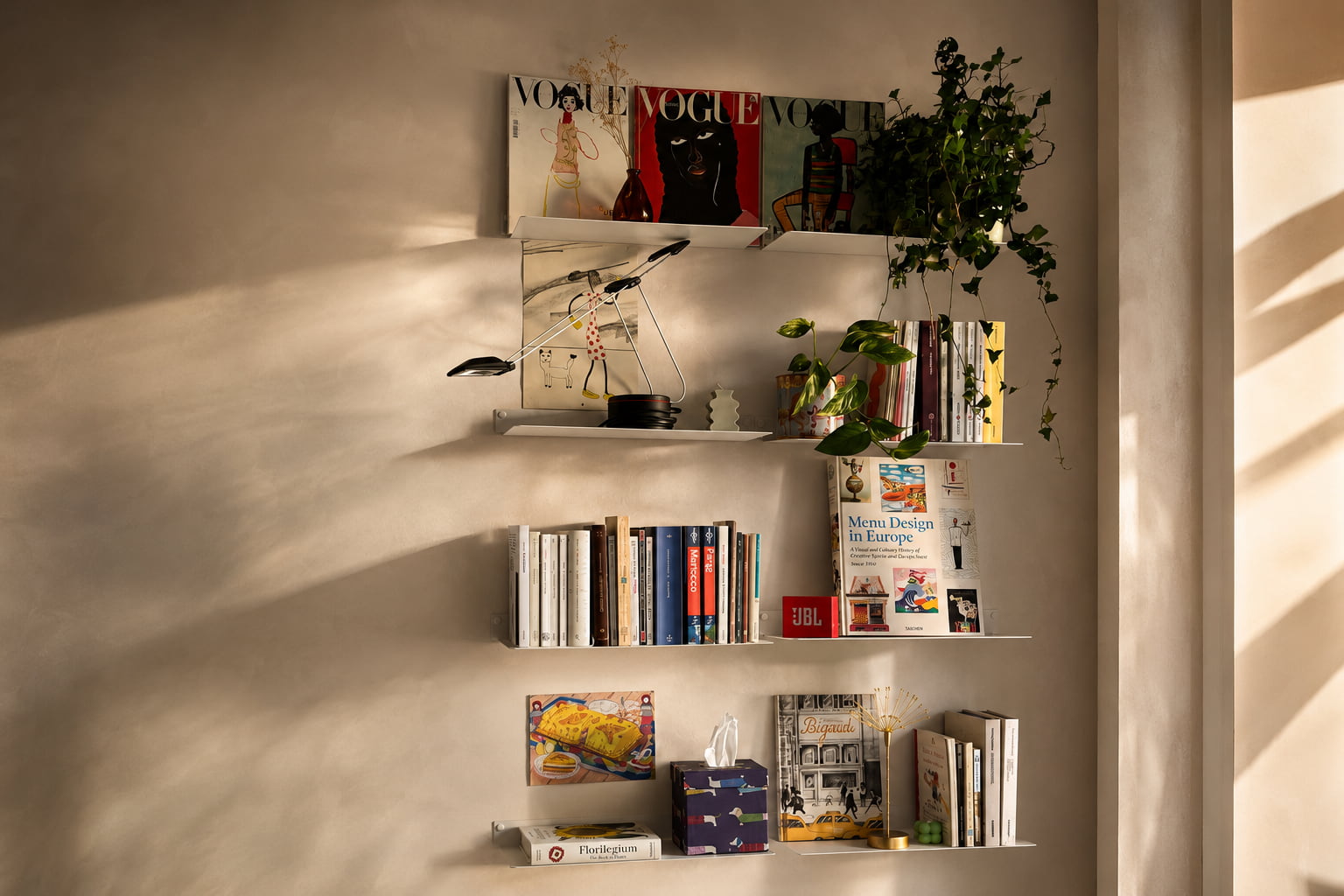 Composition de bibliothèque murale originale avec des étagères blanches décalées, accueillant livres, magazines, plantes et objets décoratifs sur plusieurs niveaux.