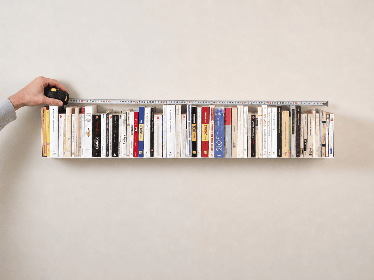Étagère à fixation invisible blanche remplie de livres, mesurée avec un mètre ruban tenu par une main, sur un mur clair au style minimaliste.