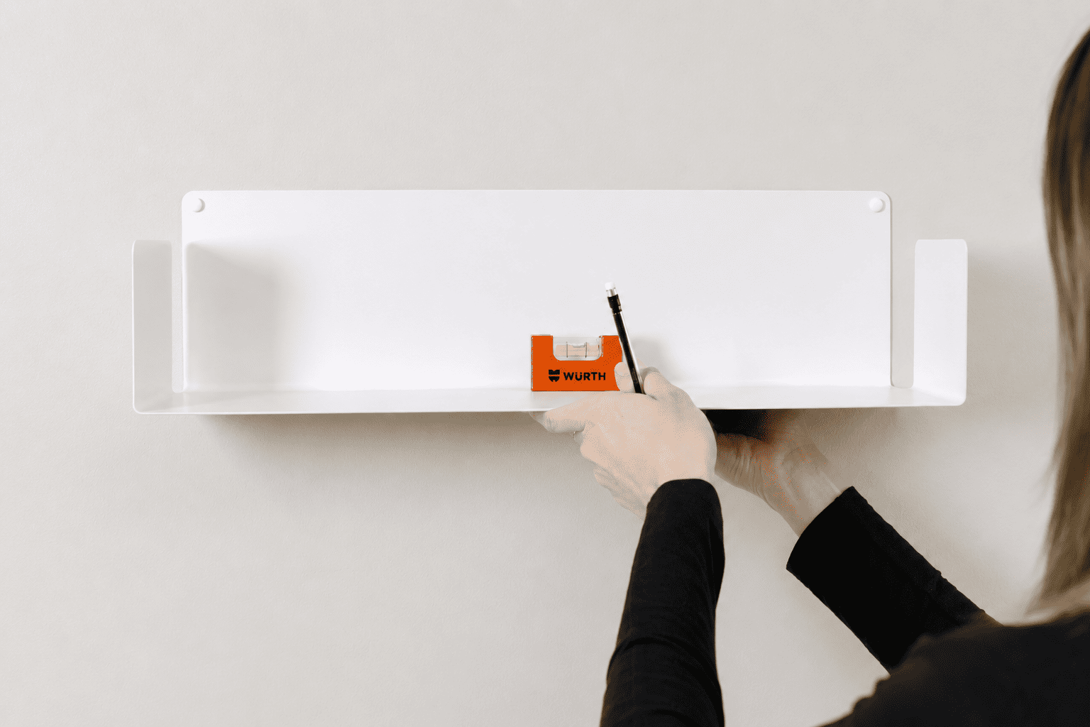 Personne utilisant un niveau à bulle pour aligner une étagère flottante blanche sur un mur clair avant la fixation, dans un intérieur minimaliste et lumineux.
