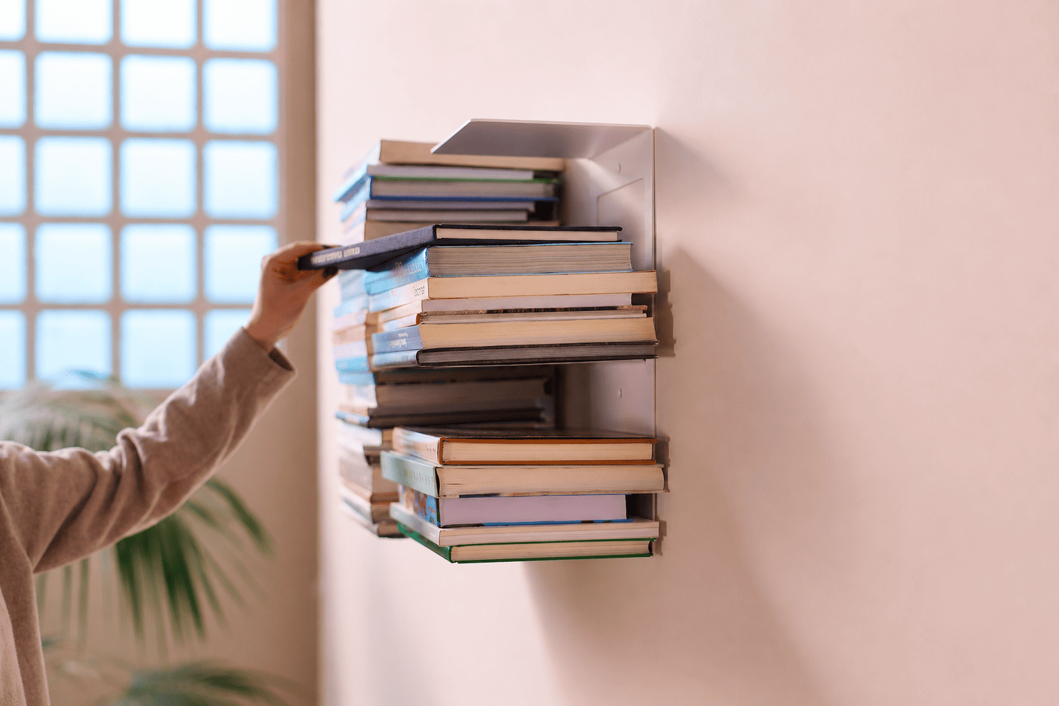 Étagère verticale en acier blanc fixée au mur, remplie de livres empilés, avec une main qui en ajoute un, dans un intérieur lumineux au style minimaliste