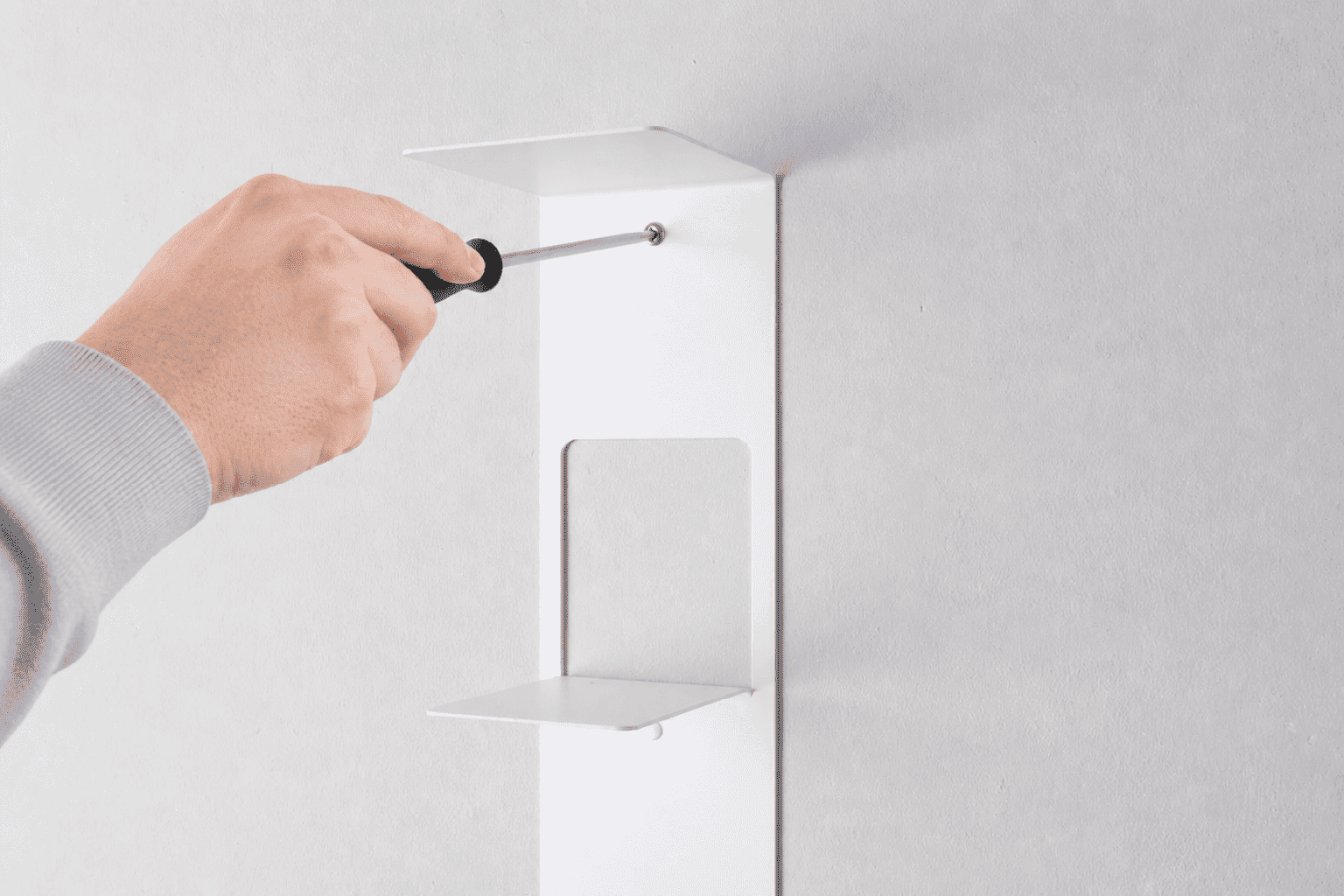 Étagère verticale fixée au mur en acier blanc en cours d’installation, avec une main tenant un tournevis pour visser le support sur un mur clair, dans un style minimaliste et soigné