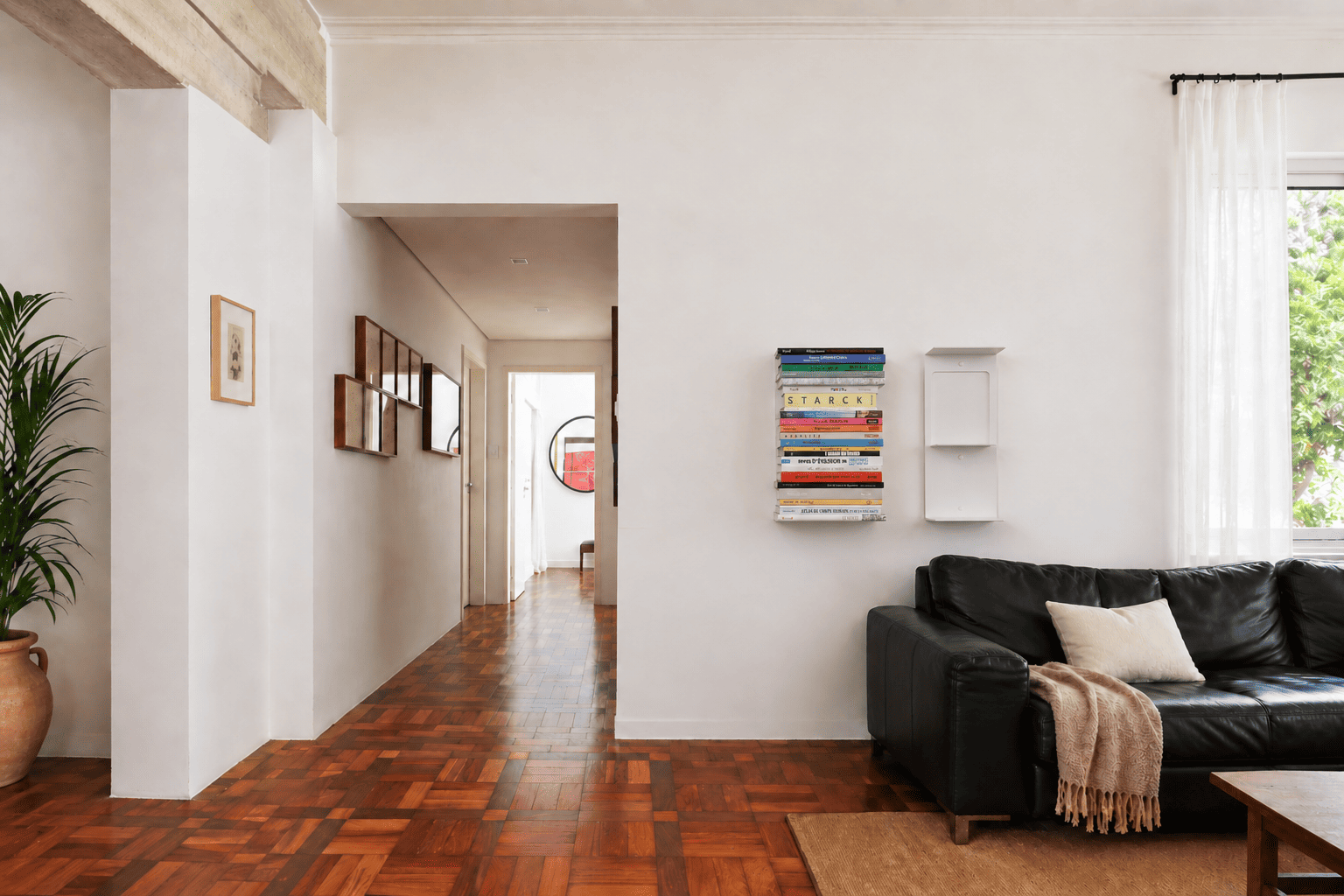 Étagère verticale solide murale en acier blanc, l’une remplie de livres colorés et l’autre vide, installées au-dessus d’un canapé en cuir noir dans un salon lumineux au parquet en bois.