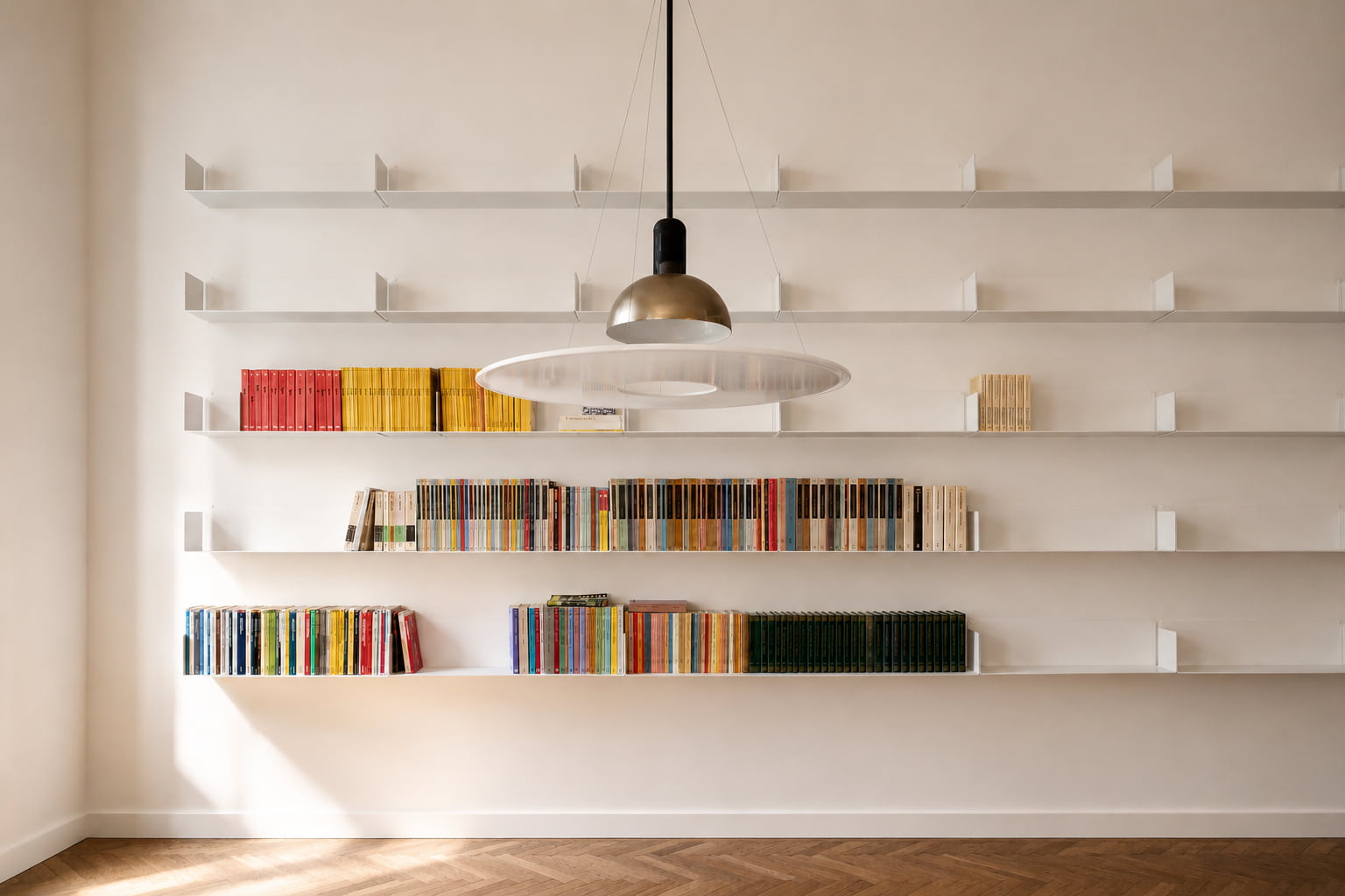 Bibliothèque murale blanche avec de longues étagères remplies de livres rangés par couleur, une idée de décoration graphique et épurée pour structurer le mur.