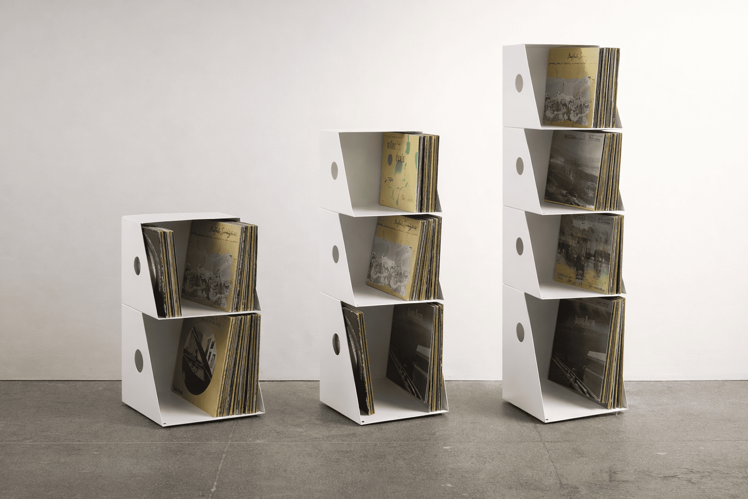 Meuble vinyle design blanc modulable en métal présenté en trois hauteurs différentes (2, 3 et 4 niveaux), avec rangement de disques vinyles dans un studio épuré sur fond neutre.