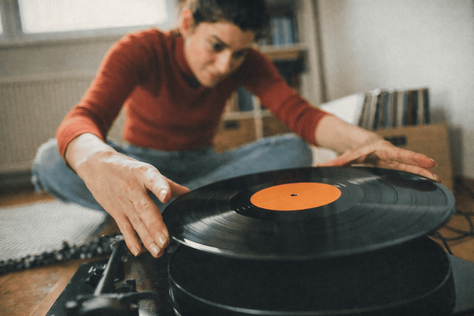 Personne manipulant un disque sur une platine vinyle, avec des étagères de rangement vinyle 33 tours visibles en arrière-plan dans une ambiance chaleureuse.